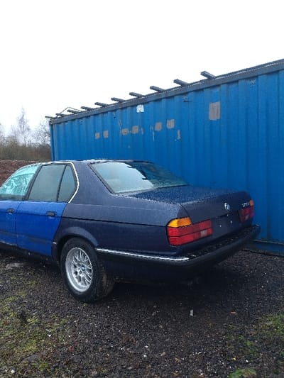 This image depicts a dark blue BMW E32 750iL sedan, viewed from its rear-left quarter, parked outdoors in what appears to be a storage or industrial area in Bitburg, Germany. The vehicle's paint appears wet, suggesting recent precipitation. Notably, the front driver's side door panel is a lighter, more vibrant blue than the rest of the car, indicating a color mismatch or repair. The rear of the BMW features a black bumper, red and amber taillight lenses, and a "750iL" badge on the right side of the trunk lid; no license plate is visible. The car is fitted with silver multi-spoke alloy wheels. The front driver's side window exhibits a frosted or heavily obscured appearance. Immediately behind the vehicle is a large, ribbed metal shipping container, painted a distinct bright blue. The container's side shows multiple faded or removed rectangular patches. Several dark, horizontal slats or beams rest along the top edge of the container. The ground beneath the vehicle is an uneven mixture of dark gravel and dirt, interspersed with small patches of green vegetation, all appearing damp. In the distant left background, sparse, bare trees or shrubs are visible against a bright, uniformly white, overcast sky. No individuals or active subjects are present in the scene.