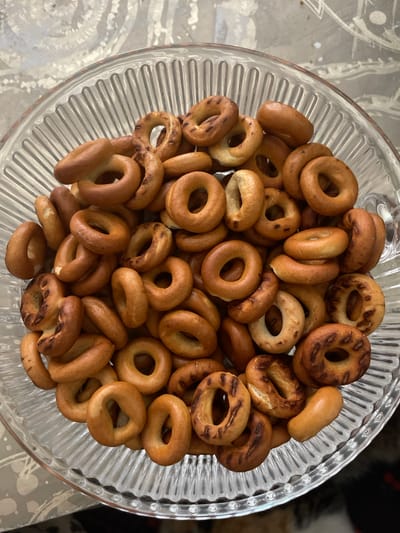 A clear glass bowl filled with small, ring-shaped cookies, commonly known as "baranki" or "sushki" in Russia, is presented from a top-down perspective. The cookies are golden brown in color, with some exhibiting darker, more toasted areas. They vary slightly in size and shape, with most being uniformly circular with a hole in the center, while a few appear more oval or elongated. They are piled generously within the fluted glass bowl, creating a textured and abundant display. The bowl itself rests on a patterned surface, possibly a tablecloth or placemat, with a soft, neutral color scheme. The background is out of focus, suggesting an indoor setting. There is no visible text in the image. The lighting is natural, casting gentle shadows and highlighting the baked goods. The overall impression is one of a simple, home-style treat, perhaps prepared for a casual gathering or as a snack. The image evokes a sense of comfort and tradition associated with Russian cuisine.