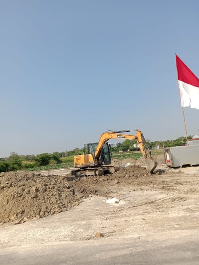 An excavator is digging on a construction site in Siwalan, Indonesia, under a clear blue sky. The excavator, a yellow SANY model, is positioned on a mound of soil, with its bucket actively engaged in moving dirt. A red and white Indonesian flag is visible flying on a tall pole to the right of the excavator. In the background, there is a line of trees and some low-lying buildings. A white refrigerated trailer is parked to the right of the excavator. The ground is a mix of loose soil, gravel, and tire tracks, indicating recent activity.