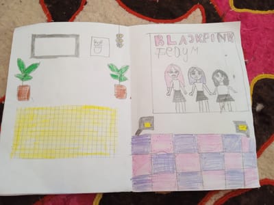 The image shows a children's drawing of what appears to be a page from a notebook or scrapbook, possibly related to the K-pop group BLACKPINK. The drawing is spread across two facing pages. The left page features a framed picture at the top with a drawing of a cat inside a smaller frame next to it. Below these are two potted plants. The lower half of the left page is filled with a grid pattern colored in yellow.
The right page has the text "BLACKPINK" written in large, colorful letters at the top, with "FE0YM" written below it in smaller letters. Below the text are three stylized drawings of young women with different hair colors (pink, purple, and black). They are holding hands. The lower portion of the right page is decorated with a checkered pattern of pink and purple squares, with some gray and yellow geometric shapes above it, possibly representing lights or speakers.
The drawing is set against a backdrop of a patterned textile, likely a rug or blanket, with colors of brown, red, and pink. The environment depicted in the drawing itself is interior, with elements like framed pictures and potted plants suggesting a room. There is no discernible connection to the city of Теңги, Kyrgyzstan, as the image contains only drawings and no real-world environmental context.
