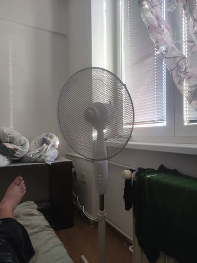 A standing fan is positioned in a room next to a window with blinds. To the left of the fan, on a bed, are two pillows with floral patterns. A person's left foot and leg are visible in the foreground, resting on a light green comforter. The fan's base and a section of its stem are visible. A dark green t-shirt and a black garment are hanging on a radiator to the right of the fan. The overall lighting suggests it is daytime, with sunlight filtering through the window. The scene is indoors and appears to be a domestic setting, likely a bedroom, in Hradec Králové, Czechia.