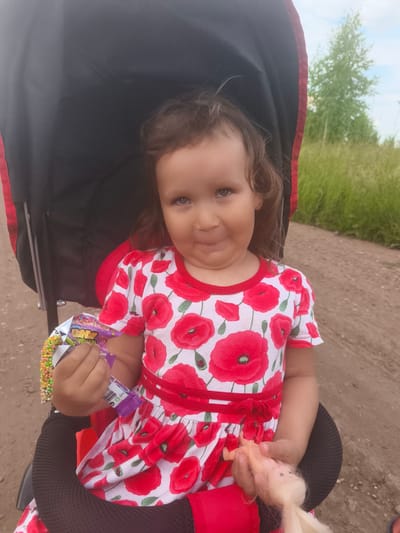 A young child is sitting in a stroller, holding a candy and a doll. The child is wearing a red and white dress with a floral pattern of red poppies. The stroller is black and red and appears to be outdoors, with a dirt path and tall green grass visible in the background. A tree is also visible in the distance. The sky is overcast, suggesting it may be a cloudy day. The child's expression is one of mild amusement. The time of day appears to be daytime. There is some visible text on the candy wrapper, but it is mostly illegible. The location is Burayevo, Russia.