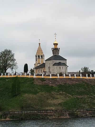 The image depicts the Church of the Presentation of the Lord in Gorodnya, Russia, situated atop a grassy hill overlooking a body of water. The church complex consists of two main structures: a taller, yellow-colored bell tower with a distinctive golden spire and a shorter, stone church with a dark roof and a golden dome topped with a cross. A yellow and white fence runs along the perimeter of the hilltop, defining the church grounds. The sky is overcast, suggesting a cloudy or rainy day. Trees are visible on the hillside and around the church grounds. A wooden fence is along the riverbank. The water is dark and choppy.