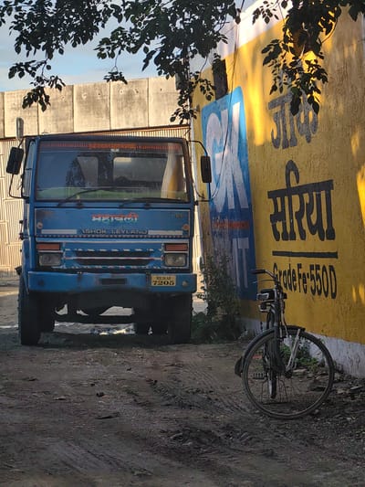 The image depicts an outdoor scene in Malkapur, India, featuring a blue Ashok Leyland truck parked on a dirt path. The truck is facing forward and appears to be the primary subject. To the right of the truck, a bicycle is parked against a yellow wall with a large blue and white graphic. The wall has dark lettering on it, including the word "सरिया" and "Grade Fe-550D." The ground is uneven and covered with dirt and small stones. Trees with dark green leaves are visible in the upper portion of the image, casting shadows on the truck and the wall. The sky is partly visible in the background, with clouds and hints of sunlight. A concrete wall is visible behind the truck. The overall impression is of a storage or industrial area.