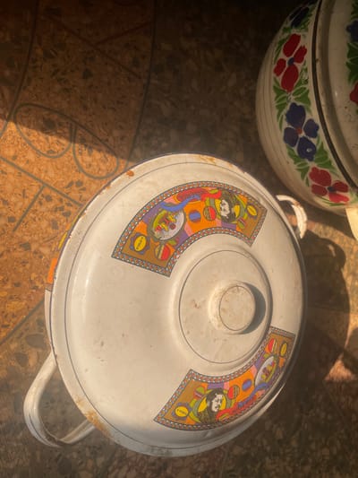 The scene depicts two enamel pots resting on a speckled, light-brown tiled floor, illuminated by direct sunlight which casts sharp shadows. In the foreground, a white enamel lidded pot, approximately circular, features multiple semicircular decals on its lid. These decals contain stylized bearded faces, the word 'NIGERIA,' 'G-STYLE,' and geometric patterns in orange, purple, green, and yellow. The pot displays significant wear, including rust spots and enamel chipping, particularly along its edges and handles. Partially visible in the upper right quadrant is a second white enamel pot, adorned with red, blue, and green floral patterns. No individuals are present. This scene is located in the city of Isiamigbo, Nigeria.
