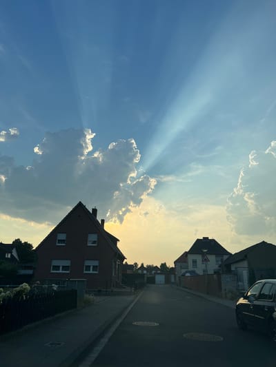 The image shows a street scene in Heinsberg, Germany, during sunset. The sun's rays are visible, piercing through the clouds, creating a dramatic effect in the sky. Several houses with brick facades and pitched roofs line the street. A dark-colored car is parked on the right side of the street. The sky is a mix of blue and warm yellow hues, indicating the approaching evening. The overall mood is peaceful and serene.