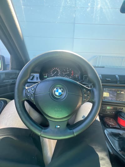 The image is a first-person perspective from inside a BMW car, likely parked in Koziegłowy, Poland, due to the distinctive exterior wall visible through the windshield. The focus is on the steering wheel and dashboard.
The steering wheel is black leather, with the BMW logo prominently displayed in the center. Buttons for car controls are visible on the steering wheel. The instrument cluster, including the speedometer and tachometer, is visible behind the steering wheel. To the right of the steering wheel, a car radio or infotainment system is partially visible.
The interior of the car appears to be a typical BMW from the late 1990s or early 2000s. The dashboard is made of black plastic and has air vents. The car's exterior is visible through the windshield, showing a large, light-colored, horizontally paneled wall that suggests an industrial building or a large garage. The sky above is bright and clear, indicating daytime.
The scene is static; the car is not in motion. There are no people clearly visible beyond the driver's legs, which are clad in light-colored shorts and black pants. The overall impression is one of a parked vehicle on a sunny day.
