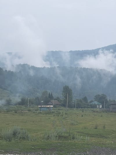The image depicts a misty rural landscape, likely in the vicinity of Ust-Kabirza, Russia. In the foreground, a grassy field stretches out, sparsely populated with small plants and bushes. A dirt path is visible, winding its way through the field. In the middle ground, several buildings are scattered amongst trees. One building appears to be a traditional wooden house with a dark roof, and next to it is a smaller, lighter-colored structure, possibly a shed or outbuilding, with a green roof. A weathered, blue metal structure with visible rust is also present. Further back, a dense forest covers the lower slopes of a mist-shrouded mountain range. The mountains themselves are largely obscured by heavy clouds and fog, creating a serene and somewhat ethereal atmosphere. The sky is overcast and a uniform pale gray. The overall impression is one of quiet, rural life, possibly early morning or a day with poor visibility due to the pervasive mist.