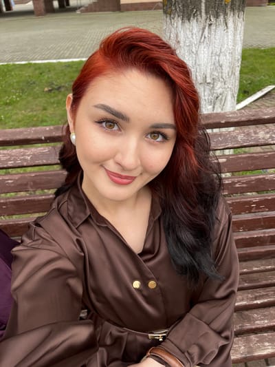 A woman with auburn and black hair is sitting on a wooden bench outdoors. She is wearing a brown satin top with gold buttons. The woman is looking at the camera and smiling. Her eye makeup is visible. She is wearing a pearl earring. The background shows a tree trunk and green grass. The photo appears to have been taken during the day, with natural lighting. The overall mood is casual and relaxed.