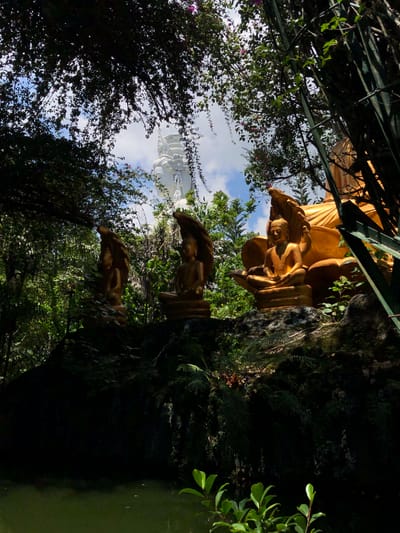 This image depicts a serene outdoor scene, likely in Nam Ban, Vietnam, featuring religious statuary amidst lush greenery. In the foreground, three golden Buddha statues are seated on a rocky outcrop. The leftmost statue is partially obscured by foliage. The central and rightmost statues are clearly visible, with the rightmost statue positioned slightly forward. Behind these figures, a very large, ornate white statue of a deity or Buddha, possibly Guanyin, rises into the sky. This immense white statue is framed by the dense, dark foliage of trees and vines, with glimpses of the blue sky and white clouds visible through the canopy. The overall impression is one of tranquility and spiritual grandeur, with nature playing a significant role in the composition.
