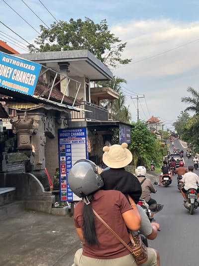The image shows a street scene in Singakerta, Indonesia. Several scooters are driving on the road. The focus is on an "Authorized Money Changer" business, whose signage is prominently displayed, listing various currencies. People are riding scooters in the foreground and background. The environment includes buildings, trees, and overcast skies.