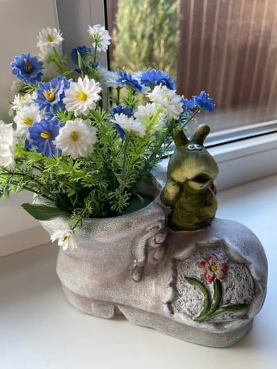 Here is a detailed description of the image:
The image shows a decorative flower arrangement in a pot shaped like a frog. The pot is grayish in color and appears to be made of stone or ceramic, with a whimsical design incorporating a frog's head and body. The frog's head is green with darker accents.
Arranged in the pot are artificial flowers in shades of white and blue, interspersed with green foliage. The arrangement is full and overflowing from the pot. The scene is lit by natural light.
The image was taken indoors, likely on a windowsill or a surface near a window, as suggested by the window frame and light filtering through. The location is identified as Лесной Пейзаж Кп (шарапово) in Russia.