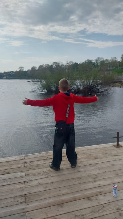 The image depicts a young male child standing on a wooden pier in Nikolayevsk-on-Amur, Russia, facing a body of water. The child, appearing to be approximately 8-12 years old, has short, light brown hair and is dressed in a red hooded sweatshirt with vertical text detailing on the back, dark baggy jeans, and dark sneakers. A small black shoulder bag is worn diagonally across his back, resting on his left hip, with a small blue accessory attached. His arms are outstretched horizontally to his sides, palms facing outward, suggesting an expansive gesture.

The pier is constructed of light brown wooden planks. To the lower right, a discarded white and blue aluminum beverage can, labeled "МИЛКИС" (Milkis) in Cyrillic, rests on the pier. A brown metal post with a horizontal crossbar is visible on the right side of the pier near the water's edge.

The body of water, likely a river or large lake, exhibits small ripples on its greyish-blue surface. The opposite bank features a dense line of green-leafed trees and bushes. Beyond the vegetation, several light-colored buildings with dark roofs are discernible, indicating a populated area. A gazebo-like structure is also visible on the distant right bank. The sky above is largely overcast with grey clouds, though patches of lighter blue sky are visible. The overall scene suggests an outdoor, daytime setting under mixed cloudy conditions.