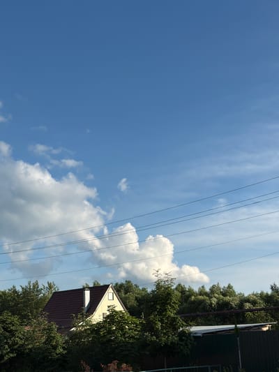 In the city of Uzyukovo, Russia, this image depicts a clear blue sky with scattered white clouds. Several power lines traverse the upper portion of the frame. In the middle ground, a house with a dark brown roof and yellow siding is visible, surrounded by lush green trees. A white chimney extends from the roof. In the foreground, more trees and a dark green fence are present, along with a low structure with a corrugated metal roof. The scene suggests a rural or suburban setting.