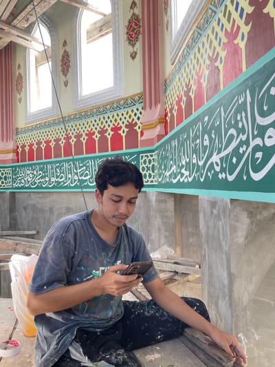 The image captures a young male seated within a mosque undergoing construction or renovation in Mns Asan, Indonesia. The male, with dark hair and a light complexion, wears a dark, paint-splattered t-shirt and pants. He is looking downwards, holding a dark-colored smartphone with both hands. He sits on a surface composed of wooden planks, with construction debris including plastic bags and a roll of tape visible nearby. The background features elaborately decorated interior walls. A prominent teal-green band displays white Arabic calligraphy, which appears to be a religious inscription. Above this band, the wall features intricate patterns, including red arched motifs and a yellow and blue geometric lattice. Further up, light-colored walls with pink-striped columns frame tall, arched windows that allow bright light to enter. Exposed wooden structural beams are visible above the windows, indicating ongoing architectural work.