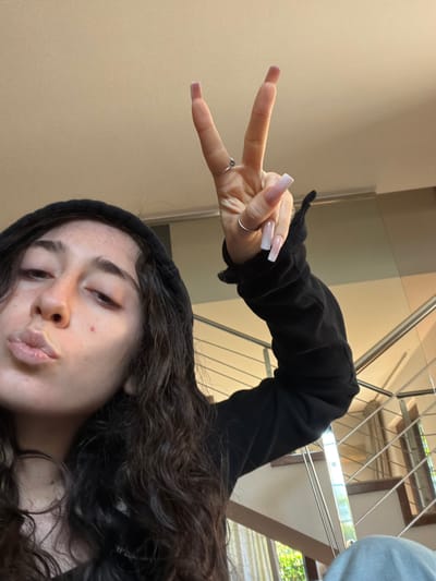 A selfie taken indoors depicts a person with long, dark, curly hair wearing a black hooded sweatshirt. The person is making a kissing face towards the camera and holding up their hand in a peace sign gesture. The person has an oval face shape, fair skin, dark eyebrows, and dark eyelashes. Their nails are long and painted with a gradient from nude to white. A silver ring is visible on their ring finger.

The background consists of a beige wall on the left and a staircase with a metal railing and wooden steps in the upper right. The location is identified as Lissone, Italy.