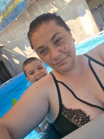 This is a selfie taken from a low angle in an outdoor swimming pool. Two people are visible: a woman in the foreground and a young boy behind her. The woman has dark hair and is wearing a black bikini with lace detailing. She has freckles on her face and chest. The boy has short brown hair and is smiling broadly, peeking over the woman's shoulder. They are both in the blue water of the pool. The background shows the concrete wall of a building, some wooden structure, and a blue and white patterned pool liner. Sunlight is casting shadows on the wall, suggesting it is daytime, likely afternoon given the angle of the shadows. The overall mood appears cheerful and relaxed, indicating a fun day at the pool. No visible text is present in the image. The location context is Kalipetrovo, Bulgaria.