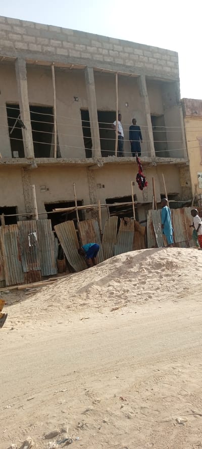 The image captures an outdoor scene in Rijiyar Zaki, Nigeria, featuring an unfinished two-story building, sandy terrain, and several individuals under bright daylight.

The building, constructed with concrete blocks, dominates the background. Its upper floor has several open rectangular frames, possibly for windows or doors, with concrete pillars and wooden supports. Two men are standing on this upper level; one in a white t-shirt and another in a dark, long garment. A bundled red garment or fabric hangs from the upper floor by a rope or wire, dangling towards the ground level. The lower floor of the building also shows open frames and is partially obscured by a temporary fence made of corrugated metal sheets.

In the foreground, a large pile of light-colored sand occupies the central and right portions of the frame. The sand shows numerous footprints and tire tracks. To the left of the sand pile, a person wearing a blue top and shorts is bending over near the corrugated metal fence. Two children are visible to the right, standing on or near the sand pile; one is dressed in a light blue garment, and the other in a light top and reddish-orange pants. The immediate foreground shows a sandy path or road, also marked with tire tracks and footprints, and scattered with some small debris.

The scene suggests an ongoing construction project or a developing area. The bright, clear sky indicates a sunny day, likely late morning or early afternoon. No discernible text is visible in the image.