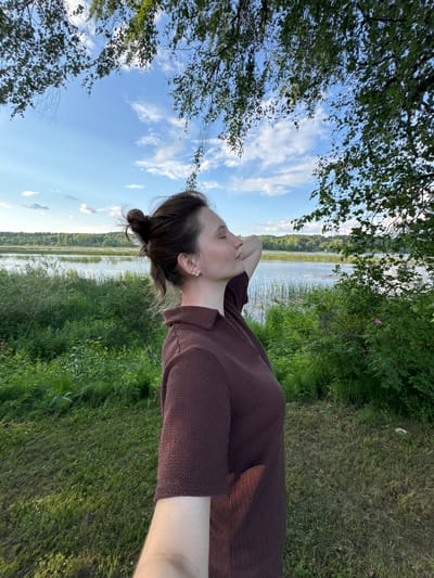 A woman with her hair pulled up in a bun stands in profile to the camera, with her head tilted back and eyes closed, as if enjoying the sun. She is wearing a brown knitted sweater with a collar and short sleeves. Her right arm is bent and resting behind her head. The background features a body of water, possibly a lake, with tall grass and reeds growing along its edge. Beyond the water, a dense forest of green trees stretches across the horizon. The sky is blue with scattered white clouds, and branches of a tree with green leaves hang into the frame from the top. The scene appears to be outdoors in a natural setting, likely a park or a scenic overlook.