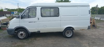 A white GAZ-2705 van is parked on a gravel lot outdoors. The van is facing left and is viewed from the side. The van is empty and appears to be stationary. The van has a sliding door on the side and a window in the front passenger area. The lot appears to be a storage yard, with concrete blocks and metal fencing visible in the background. The sky is overcast, and there are trees and buildings in the distance. There are no people or animals visible in the image. The van has rust spots on the lower body panels and the rear bumper is missing a section.