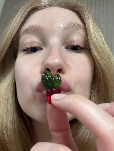 A young, blonde woman is holding a cactus-shaped gummy candy between her lips. Her face is fair and she has long eyelashes. The scene is indoors, the background is indistinct. There is no indication of the location being Yuzhny, Russia.