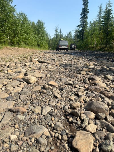 The image depicts a rocky dirt road in Talnakh, Russia, leading into a forested area. Two dark-colored off-road vehicles, possibly SUVs, are visible in the distance on the road. The foreground is dominated by a close-up view of the rough, uneven terrain composed of various sizes of grey and brown stones and pebbles. The surrounding environment consists of dense green deciduous and coniferous trees, with a clear blue sky overhead. The scene appears to be a remote, natural landscape.