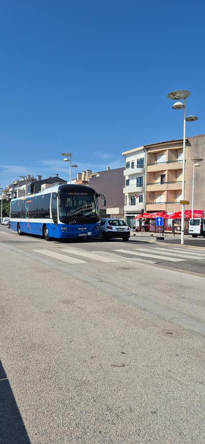 A blue and white MAN bus, with "unir" branding and the route number "3301 RU AGUÇADOURA" displayed, is stopped at a pedestrian crossing on a sunny day in Vila do Conde, Portugal. The bus is positioned on the right side of the road, facing forward. To the right of the bus, a white Peugeot 207 hatchback is also stopped at the crossing. In the background, there are multi-story buildings with balconies, and streetlights with distinctive circular shades are visible. A yellow sign with the word "Desvio" (Detour) and an arrow points to the right. A white van is parked further down the street. The road is paved and a pedestrian crossing is marked with white stripes. The sky is clear and blue. No people are clearly visible in action, though there might be people inside the shops or buildings. The scene depicts a typical urban street with public transportation and vehicular traffic.
