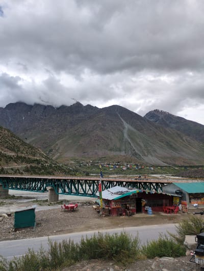 The image depicts a roadside scene in Darcha, India, with a focus on a small cluster of roadside eateries and shops. A long, green truss bridge spans a river in the background, with steep, rocky mountains rising behind it. The sky is overcast with heavy clouds, suggesting a potential for rain or a generally gloomy atmosphere.
In the foreground, a paved road runs horizontally across the bottom of the image. A motorcycle is parked on the right side, partially visible. To the left of the road, several small shops and food stalls are lined up. One prominent red building with a green roof has signs indicating it is a food corner. A person, possibly an employee or customer, is seated on a chair outside this establishment. Red tables and chairs are set up outside some of the shops, suggesting outdoor seating for customers. A small, green shed is also present to the left.
Further back, the truss bridge is a dominant feature, its green structure contrasting with the muted tones of the landscape. The river flows beneath it, its banks appearing rocky and sparsely vegetated. On the opposite side of the river, a small village or settlement is visible nestled at the base of the mountains, with colorful prayer flags strung across the landscape. The mountains themselves are rugged and imposing, with patches of vegetation and scree slopes. Some snow can be seen on the higher peaks. The overall impression is one of a remote, mountainous region with essential services catering to travelers.