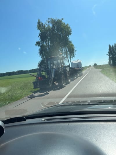 The image shows a blue tractor with a trailer carrying a large white object, possibly a bale of hay or a large package, driving on a paved road. The tractor is on the right side of the road, moving away from the viewer. The road is bordered by a green grassy field on the left and more grass and trees on the right. The sky is clear and blue. The image appears to be taken from inside a vehicle, with the dashboard visible in the foreground. The location is identified as Tuksy, Russia.