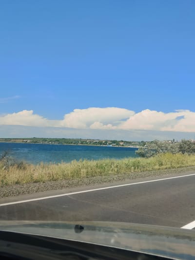 The image shows a scenic view of a body of water with a town in the distance, seen from a road. The sky is blue with fluffy white clouds. The foreground features a paved road with a white line marking the edge. To the left of the road, there is a strip of gravel and then tall, dry grass. Beyond the grass is the blue water, which appears to be a bay or lake. In the background, across the water, is a line of buildings and trees, indicating a town or village. The overall environment suggests a warm, clear day in a coastal or lakeside area. It is a still scene, with no people or animals visible, and no apparent action taking place. The perspective suggests the photo was taken from a moving vehicle.