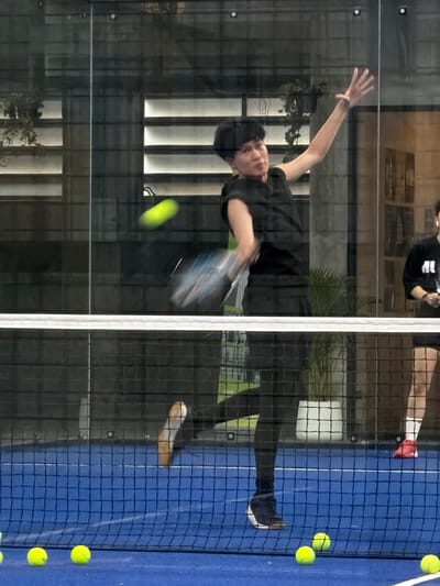 A person is playing padel on a blue court indoors at a facility in Ban Buak Khrok North, Thailand. The person is wearing a black sleeveless shirt and black leggings, and is in mid-action, striking a neon yellow ball with a padel racket. The racket is blurred due to motion, as is the ball. The player's left arm is extended upwards and to the right, with the hand open. The court has a white net dividing it, and several neon yellow padel balls are visible on the court floor in the foreground. The background shows a glass wall with reflections of plants and what appear to be blinds or louvers on another wall. Another person can be partially seen on the right side of the frame, wearing red shoes and white socks. The lighting is artificial.
