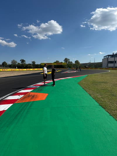 The image displays a section of the Hungaroring race circuit in Mogyoród, Hungary, observed on a clear, sunny day with a blue sky and scattered cumulus clouds. The foreground shows a dark asphalt racetrack curving to the right, bordered by a painted red and white curb. Adjacent to the curb is a wide, bright green painted asphalt runoff area. An orange-painted, roughly trapezoidal section is embedded within the green runoff, exhibiting black tire marks extending onto the green surface.

Two adult males are standing on the green runoff area near the curb in the foreground. The individual on the left wears a white t-shirt and dark shorts, facing the individual on the right who wears a dark t-shirt and dark trousers. Their posture suggests they are engaged in conversation or focused observation of the track. Further down the track, along the curve, approximately four to five additional indistinct figures are visible standing on the green runoff.

The track's perimeter is lined with yellow safety barriers displaying "PIRELLI" branding. A large yellow gantry, also bearing "PIRELLI," spans the track in the middle distance. To the far right, a multi-story modern building with a dark upper section, resembling a pit building or control tower, is visible. Trees line the horizon in the background, and a tall, thin pylon or tower is discernible against the sky. Shadows cast by the individuals and objects indicate bright, direct sunlight.