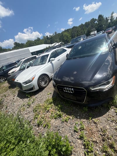 This is a ground-level, wide-angle shot of a car lot in the Village of Johnson City, United States of America, on a sunny day with scattered clouds. In the foreground, a dark grey Audi sedan is parked, facing towards the left of the frame. The car's grille, headlights, and hood are visible, reflecting the sky. Behind the Audi, several other vehicles are lined up, including a white sedan with dark accents on its hood, and further back, a dark SUV. In the background, a long, white, corrugated metal structure with a green section is visible, likely part of a building or storage area. A dense line of green trees forms a backdrop to the scene, with a clear blue sky overhead. The ground is covered in gravel and patches of green vegetation.