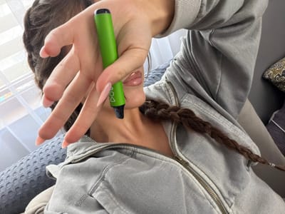 The image shows a close-up view of a person's hand holding a green vape pen in front of their face. The person is wearing a gray hooded sweatshirt. Their hair is dark brown and braided. The background is blurred, but appears to be an indoor setting with a window and curtains. The image does not provide any information about the city of Zmeitsa, Bulgaria.