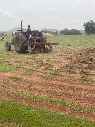 A tractor is tilling a field in Durumi, Nigeria. A person is driving the tractor. The field is partially tilled, with a dirt path to the right. Green grass is growing in the field. There are trees and hills in the distance. The sky is overcast.