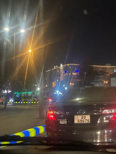 The image depicts a nighttime urban street scene in Dabouq, Jordan, viewed from inside a vehicle. The foreground shows the top of a dashboard or windshield frame. Immediately ahead, the rear of a black sedan, likely a Toyota, is visible with its red taillights illuminated. A Jordanian license plate, bearing the numbers "20 59676," is centrally affixed to the trunk. A white sticker with "SELLERS-SEXTON" text is present on the lower-left portion of the trunk lid. To the left of the black sedan, a yellow and blue striped road barrier extends horizontally. The background features multiple bright streetlights, including a prominent orange-yellow one, which create significant lens flares across the image. Multi-story buildings are visible beyond the street, adorned with string lights along their facades and exhibiting some blue-lit windows. Other vehicles are present on the road, indicated by their illuminated headlights and taillights, suggesting traffic conditions. The sky is dark, consistent with nighttime.