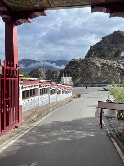 The image depicts a view through an arched entrance decorated with murals, framing a scene in Thiksey, India. In the foreground, a red metal gate stands to the left, partially open and secured with a padlock. Beyond the gate, a long wall with white and red trim lines a paved walkway. This wall features numerous prayer wheels, each adorned with intricate carvings, arranged in a row.
The paved walkway leads to a large, open asphalt area that appears to be a courtyard or parking lot. In the middle ground, several white stupas with golden spires are visible, set against a backdrop of rugged, mountainous terrain. A white van is parked to the right of the stupas. A solitary figure, dressed in dark clothing, stands in the courtyard, and to the far right, a small green bush grows near a red-framed signpost. The sky above is overcast with dramatic clouds, and layers of mountains recede into the distance. The overall impression is a serene and spiritual landscape characteristic of the region.