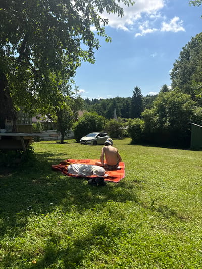 The image depicts a sunny day in a rural backyard setting. In the foreground, a person is lying on an orange blanket on a grassy lawn. The person is wearing patterned swim trunks and a hat. Another blanket or pillow is partially visible beside them. In the middle ground, a light-colored car is parked on a gravel area. Beyond the car, there are trees, a fence, and houses. The background consists of a dense line of trees and hills. The overall atmosphere is relaxed and summery, with the presence of a residential area suggesting a private property. The image appears to be taken in a location that is part of the Гаранскі сельскі Савет in Belarus, indicated by the context provided.