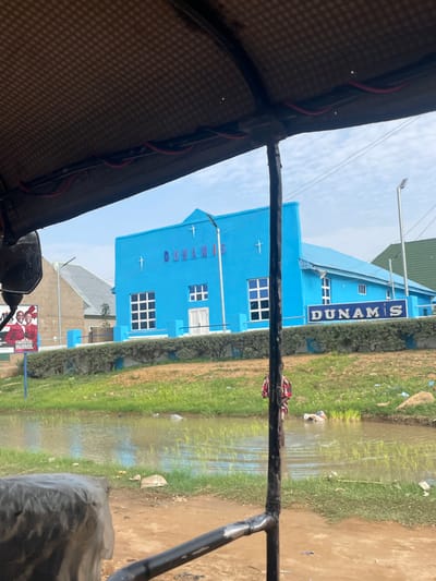The image, taken in Farei, Nigeria, shows a bright blue building with the text "DUNAMIS" prominently displayed on its facade. The building appears to be a place of worship or a community center. Several windows, some with a grid pattern, are visible on the building's exterior. In front of the building, there is a body of water, possibly a flooded area or a natural water source, with green grass growing along its edges. Some debris is scattered on the ground and in the water, suggesting a potentially untidy environment. The image is captured from the perspective of someone inside a vehicle, likely a tuk-tuk, as indicated by the visible brown patterned fabric of a canopy and parts of the vehicle's structure. Red wires are also visible running along the canopy's edge. The sky is overcast with a bright, diffused light, suggesting it could be daytime, perhaps late morning or afternoon. The overall impression is of a scene in a developing area, with a mix of structures and natural elements, hinting at everyday life in a Nigerian town.