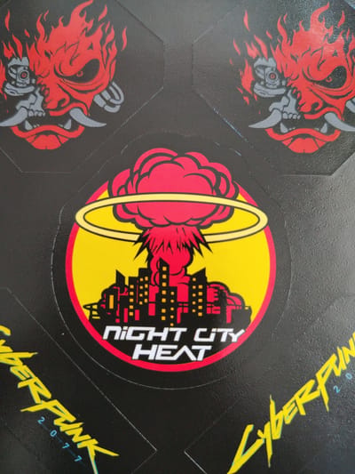 The image displays a close-up view of several stickers, likely part of a collection or a product packaging. The stickers are predominantly black with vibrant graphic designs.
One prominent sticker features a circular emblem with a yellow outer ring and a bright yellow background. Inside this ring, a red, cloud-like explosion is depicted, with a stylized yellow ring encircling the top portion of the explosion. To the right of the explosion, a cityscape is illustrated in red and yellow, with numerous rectangular windows suggested by yellow dots and lines. The text "NIGHT CITY HEAT" is printed vertically in white on the cityscape.
Another set of stickers showcases a recurring motif of a demonic or monstrous head with flowing red flames emanating from it. The head is rendered in red and gray, with sharp horns and glowing red eyes. The texture of the stickers appears to be somewhat glossy, with a subtle embossed pattern on the black background.
The overall aesthetic of the stickers suggests a theme related to a video game or a similar form of media, possibly "Cyberpunk 2077" given the visible text "CYBERPUNK 2 0 7 7" in a distinctive yellow font along the bottom right of the image. The colors are bold and high-contrast, with a clear emphasis on red, yellow, and black. The image does not provide information about the time of day, weather, or a specific emotional tone beyond the aggressive imagery. The location context of Kolomyia, Ukraine is external information and not derivable from the image itself.