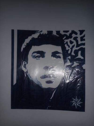The image shows a black and white drawing of a man's face on a textured surface, possibly plastic sheeting. The drawing is in a stark, high-contrast style, with dark areas representing shadows and outlines, and lighter areas for the skin and highlights. The man has dark hair, a mustache, and his eyes are looking forward. There is a decorative pattern on the right side of the drawing, and a star-like symbol in the lower right corner. The artwork is affixed to a plain, light-colored wall. There is no visible indication of the specific location within the city of Gulbag, Uzbekistan.