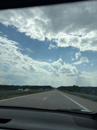 The image shows a view from a car driving on a highway. The sky is partially cloudy with blue sky visible between the clouds. There are trees and greenery on either side of the highway, with some cars driving in the distance. The highway is paved and has lane markings. The weather appears to be clear. The location is specified as Czerna, Poland.