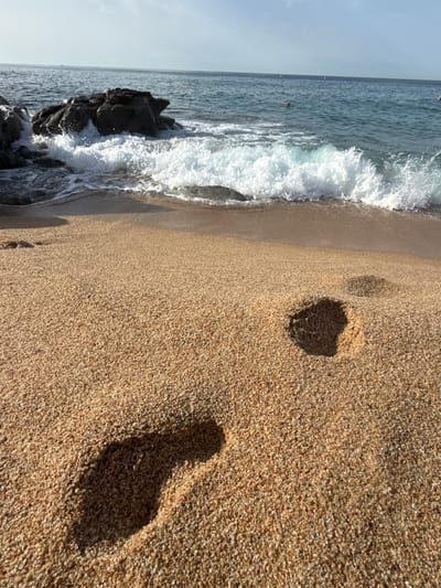 A close-up low-angle shot of a sandy beach shows two footprints pressed into the wet sand near the shoreline of the Mediterranean Sea. The footprints are in the foreground, with one deeper than the other. The sea is a deep blue with white waves crashing onto the shore. Rocks are visible in the water to the left, and the horizon stretches across the top of the frame. The sky is a pale blue with a few scattered clouds. The scene appears to be set in Lloret de Mar, Spain, a popular coastal town on the Costa Brava.