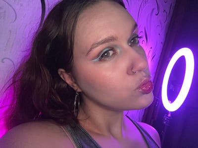 A young woman with dark hair and bold blue eyeliner is looking into the camera. She has a nose ring and large hoop earrings. She is wearing a denim top. The background is a purple wall with a swirling pattern. A bright white ring light is to her right. The image is taken from the chest up. The location is not specified in Bishkek, Kyrgyzstan.