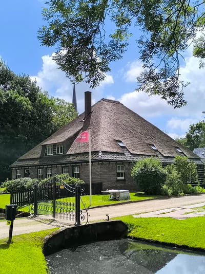 This image captures a serene rural scene in Oudorp, Netherlands, featuring a distinctive building with a large, steep thatched roof. The structure has dark wooden siding and numerous white-framed windows, including dormer windows on the roof.
In the foreground, a calm body of water, likely a pond or canal, reflects the sky. A well-maintained green lawn borders a gravel path that leads to an ornate black metal gate. A small green box on a post, marked with the number '9', stands beside the path near the water.
A red flag with white, mirrored text flies from a pole in front of the building, suggesting a local business or activity. While the exact reversed text is difficult to decipher fully, it appears to indicate something like "Ontdek Boerderij" (Discover the Farm) or related to farm activities or tours, common in such rural settings.
The property is enveloped by lush green trees, with their branches partly framing the bright blue sky dotted with scattered white clouds. In the background, the slender, dark spire of a church or similar building can be seen rising above the treeline.
The scene depicts a clear, sunny day, likely midday to early afternoon, with no people visible, contributing to a peaceful and tranquil atmosphere. The architectural style and natural surroundings evoke a sense of traditional Dutch countryside charm.