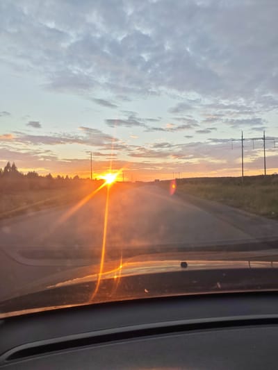 The image captures a car's view of a road at sunset, with the sun low on the horizon and casting bright rays. The location is identified as Krasnoe Pole, Russia. The foreground shows the dashboard of a car. Beyond the road, there are fields with scattered trees and power lines silhouetted against the colorful sky. The clouds are a mix of gray and soft pink, indicative of twilight. The road appears to be a two-lane asphalt highway with some traffic visible in the distance. The overall scene depicts a serene, late-day journey through a rural or semi-rural landscape.