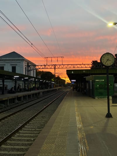 The image captures a view of a train station platform in Monte Grande, Argentina, at what appears to be dusk or dawn, judging by the pink and orange-hued sky. A clock with a white face is visible on a pole, displaying the time around 4:53. Overhead electrical wires and support structures are present. The platform surface is a combination of textured tiles. Train tracks run alongside the platform edge. A building with an angled roof and partial awning is visible at the bottom of the frame. People are waiting at the platform.