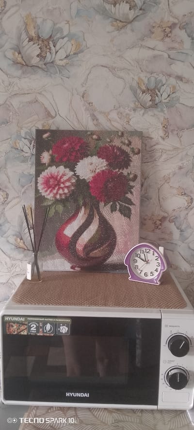 In the city of Borovka, Russia, a white Hyundai microwave oven is situated on a surface, possibly a table or counter. On top of the microwave, a woven mat is placed, and on this mat, a diamond painting of a floral arrangement in a vase is displayed. To the left of the painting, a glass bottle with dark reeds, likely an aroma diffuser, stands. To the right of the painting, a small purple house-shaped alarm clock is positioned. The background features patterned wallpaper with floral motifs. The lower portion of the image shows the front of the microwave oven, with its door and control dials visible. The brand name "HYUNDAI" is present on both the appliance and the wallpaper. A watermark from "TECNO SPARK 10c" is visible in the bottom left corner. No people are present in the scene.