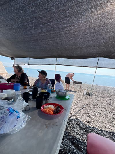 An outdoor barbecue is taking place on a rocky beach, presumably in the city of Karashygan, Kazakhstan, with the Caspian Sea visible in the background. A group of four people is present. Two women are seated at a table under a dark mesh canopy, one with a bottle of beer and the other with a can of what appears to be Beks Energy Drink. A child wearing a red and white bandana is also seated at the table. A man, shirtless and wearing black shorts and a cap, is standing and grilling food on a barbecue. On the table, there are various items including plastic cups, metal bowls, and a red plate with food. A portion of a pink object, possibly a seating cushion, is visible in the lower right corner of the image. The environment consists of a pebble beach and calm blue water under a clear sky.