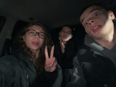 Three individuals are present in the image. On the left, a male subject with short hair is looking up. In the center, another individual is visible, with only the face and upper body visible. On the right, a female subject with curly hair and glasses is sticking out her tongue and making a peace sign with her fingers. The subjects appear to be in a dimly lit environment, potentially a car interior. There are no visible landmarks or details to confirm the location as Karavelovo, Bulgaria.